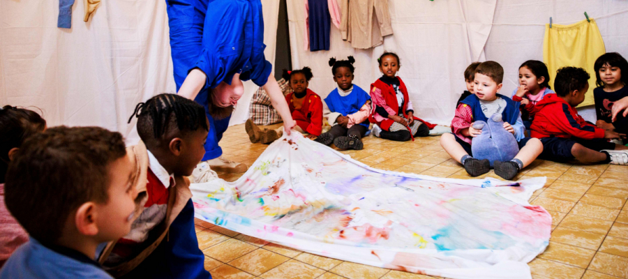 Kinderen zitten op de vloer in een klas en kijken naar twee volwassenen waarvan de ene de andere ondersteboven houdt.. 