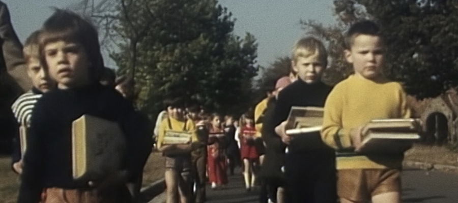 Still uit de documentaire Paradigma. Schoolkinderen wandelen in twee rijen over een weg met boeken in de hand of onder de arm.