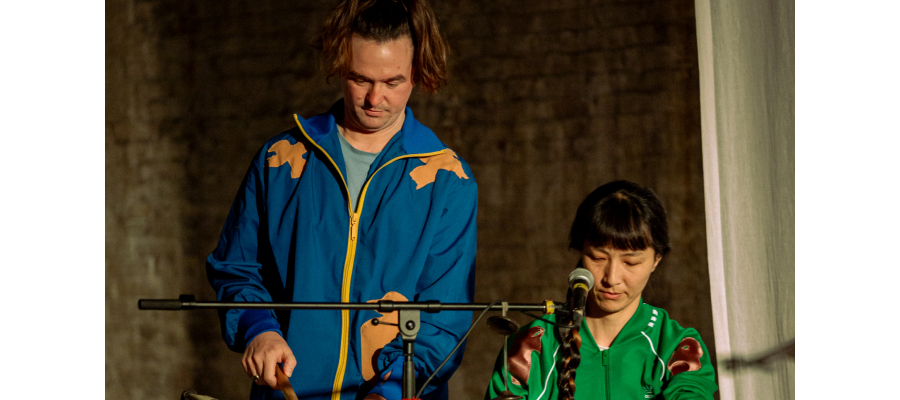 Twee muzikanten. Eentje slaat rechtopstaand op een grote trommel. De andere zit achter een Chinees muziekinstrument, wellicht een guzheng.