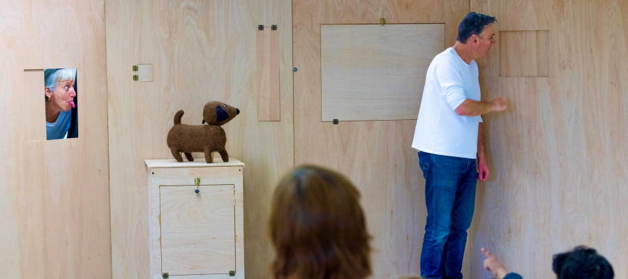 Het publiek kijkt naar een houten wand. Een vrouw staat erachter en steekt haar hoofd door een gat in de wand. Ze steekt haar tong uit naar een man die voor de wand staat. Hij kijkt een andere kant uit. Op het toneel staat ook een kastje met daarop een sp