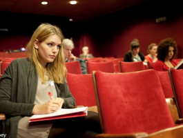 jonge vrouw zit samen met andere mensen in een auditorium of zaal en neemt nota's